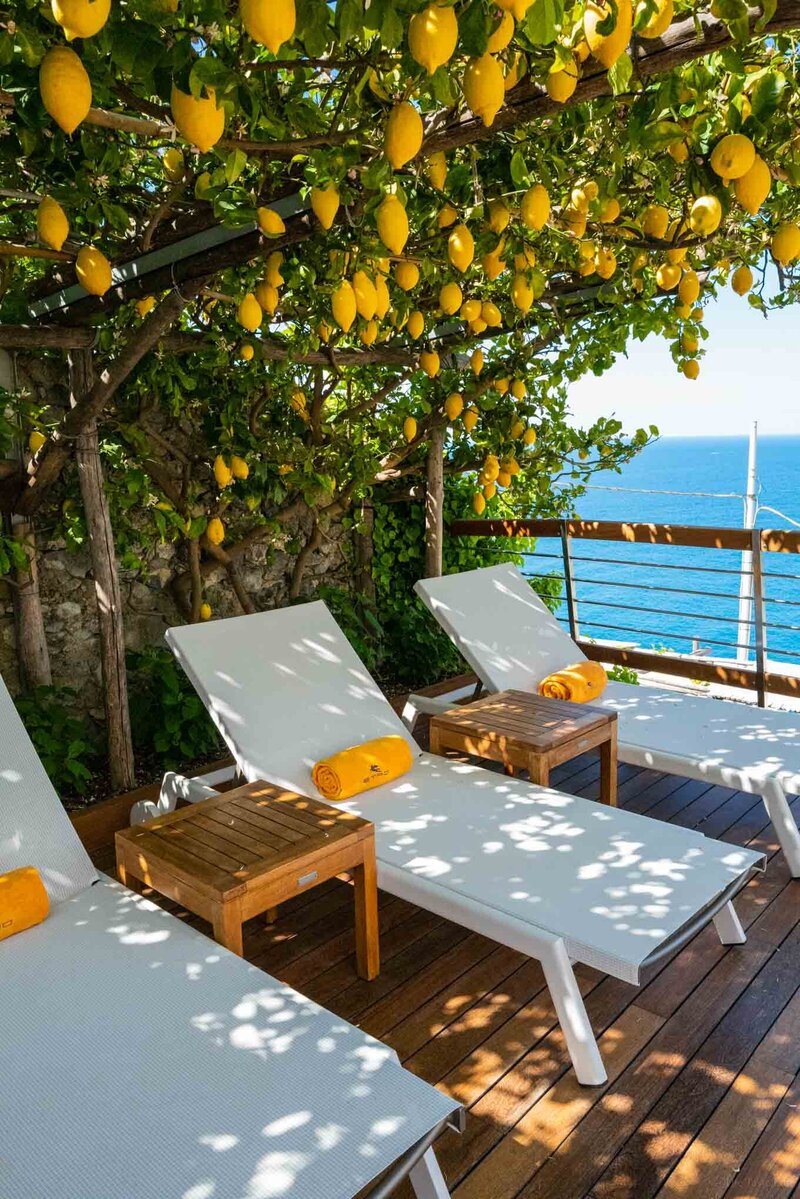 Sunlit terrace on Italy’s Amalfi Coast with lounge chairs under a canopy of ripe lemons, overlooking the sparkling blue sea and wooden deck shaded by greenery.