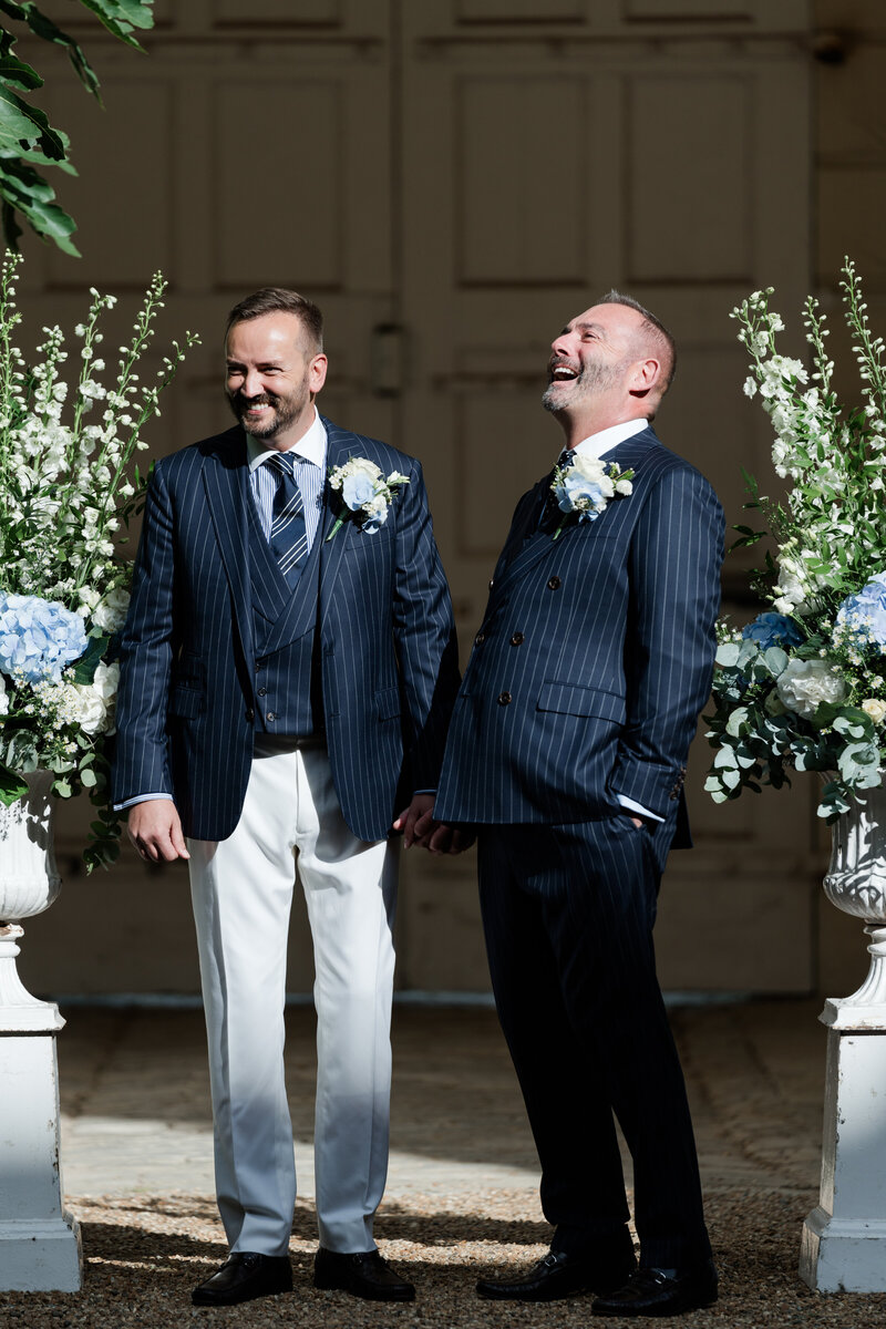Two grooms in navy pinstripe Ralph Lauren suits standing between two urns of blue hydrangea flowers laugh during their same sex wedding ceremony at Firle Place