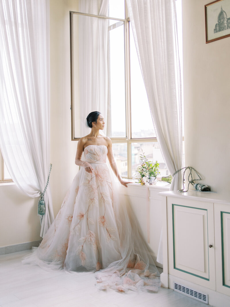 Bride stands in pink floral gown looking out the window towards the Duomo in Florence, Italy for wedding