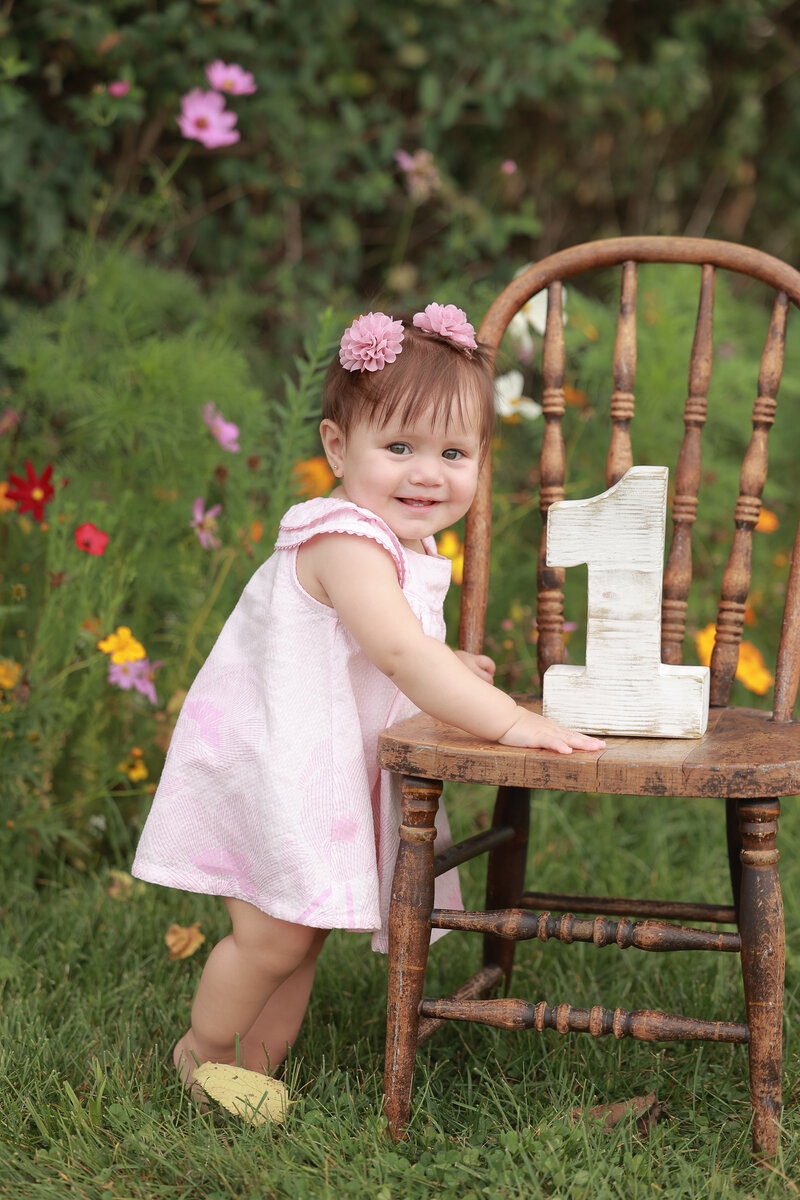 Lehigh Valley Cake Smash Photographer first birthday girl standing by a wooden chair outside with greenery and wildflowers behind her and one year old girl is in a light pink dress with pink flower hair clips