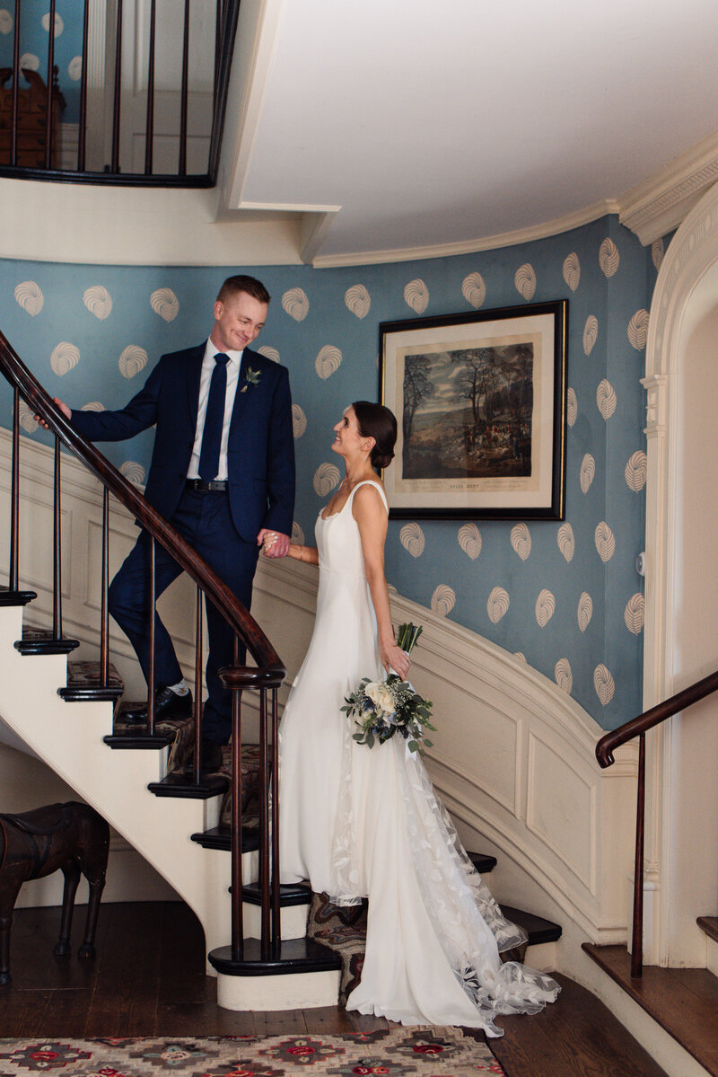 Bride follows groom up spiraling staircase at The Brick House in Shelburne, VT. Alexandra Grecco gown train and wedding flowers by A Schoolhouse Garden.