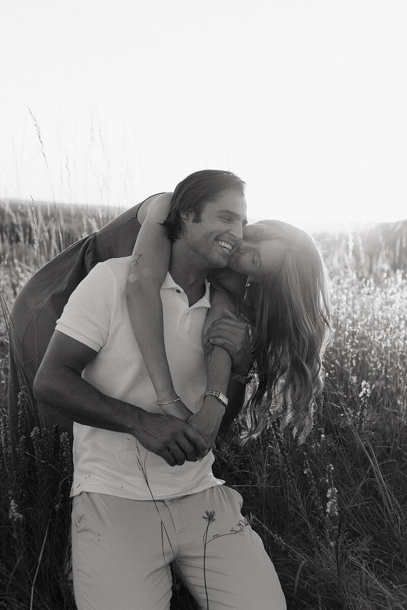 Engagement woman leans over man's back to kiss him on the cheek