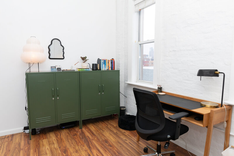 Wooden desk with black rolling chair facing two windows, green office cabinet with books atop  in an office with wood floors at Java Studios.
