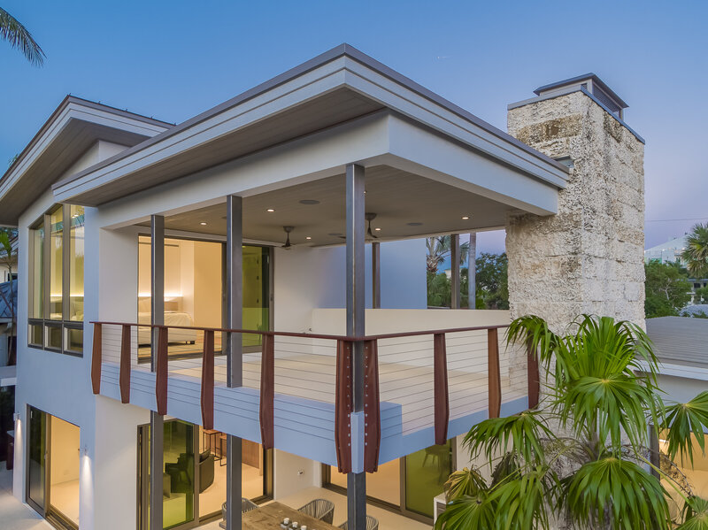 Large second story balcony on a large stone house. There is a modern brown and metal wire railing, metal beams, poured white cement floor, and a flat roof above. It is in a topical location taken at dusk, the lights are on in the house.