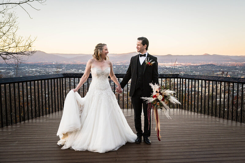 A couple on their elopement day sit on a park bench on top of a hill with their hiking backpacks. The couple wrap their arms around one another and face the view of the Blue Ridge Mountains from Heritage Park in Blacksburg, Virginia.