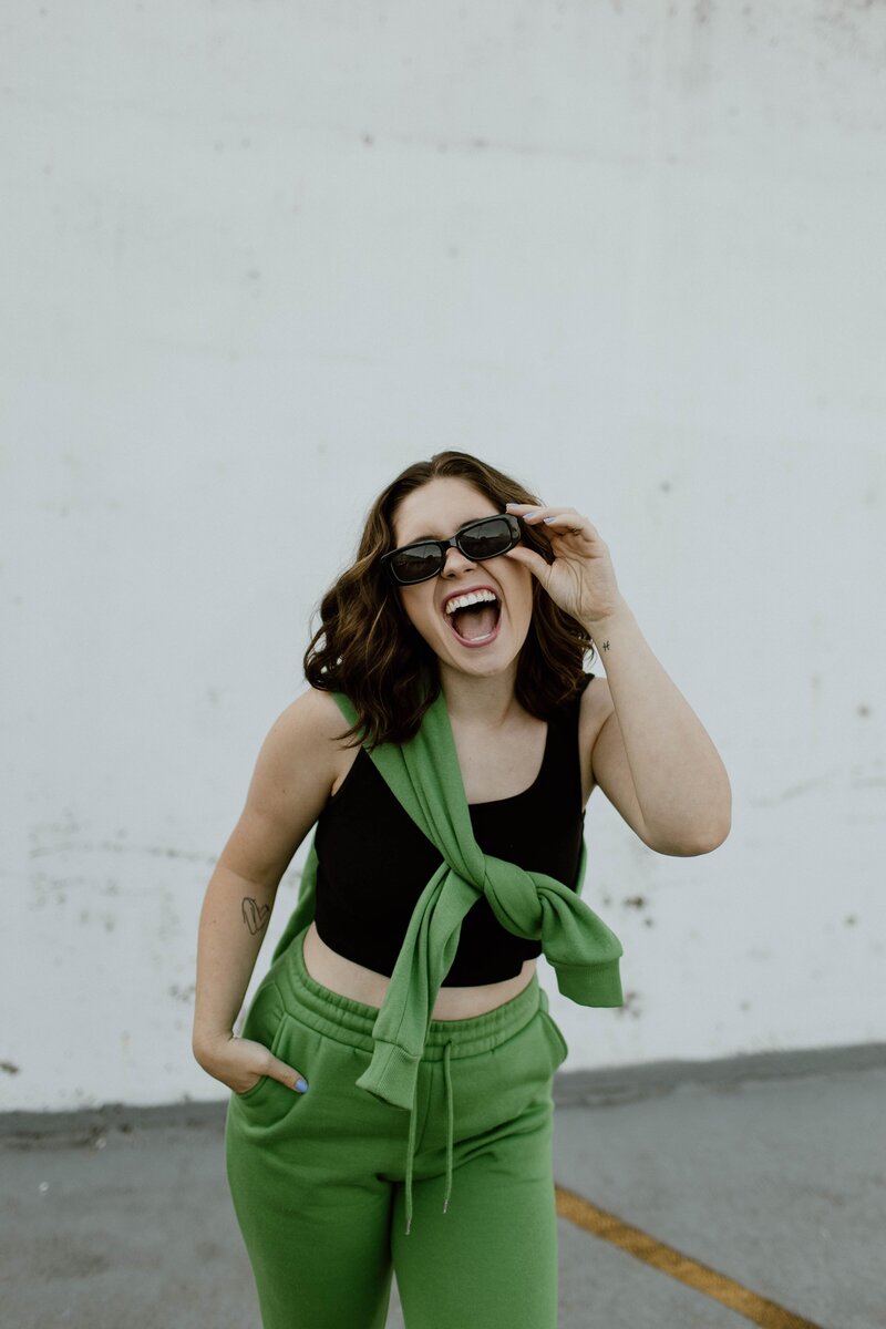 Woman laughing and holding sunglasses with one hand while wearing a black crop top, green sweatpants, and a matching green sweatshirt tied around her shoulders, standing in front of a light concrete wall.