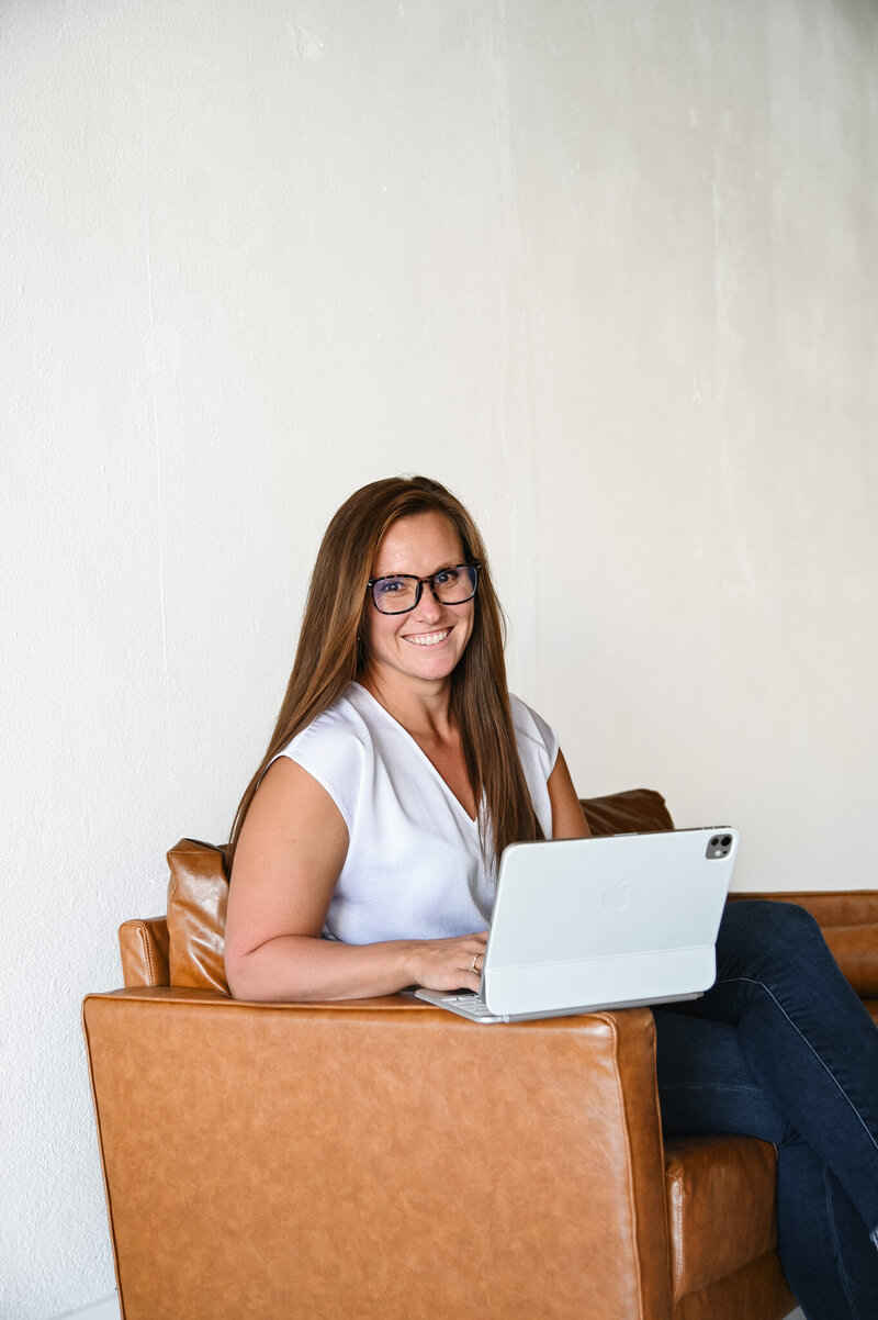 Hannah Short sitting on a leather chair with a laptop beside text answering common therapy questions about first sessions, insurance, and sliding scale options at Rooted & Nourished Psychotherapy