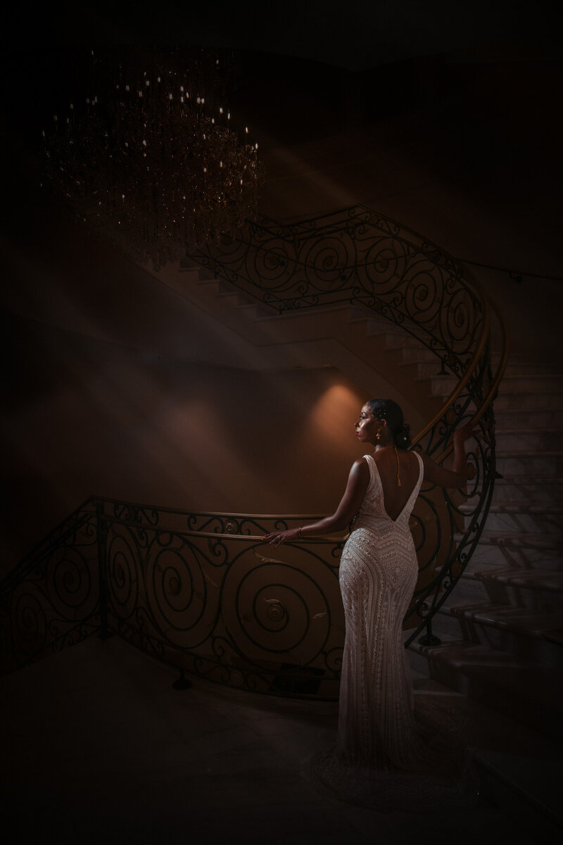 Light shining through on a bride standing at the bottom of a grand staircase.