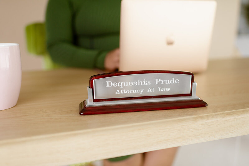 Close-up of a wooden desk nameplate with 'Dequeshia Prude, Attorney At Law' engraved, partially obscured by a laptop edge.