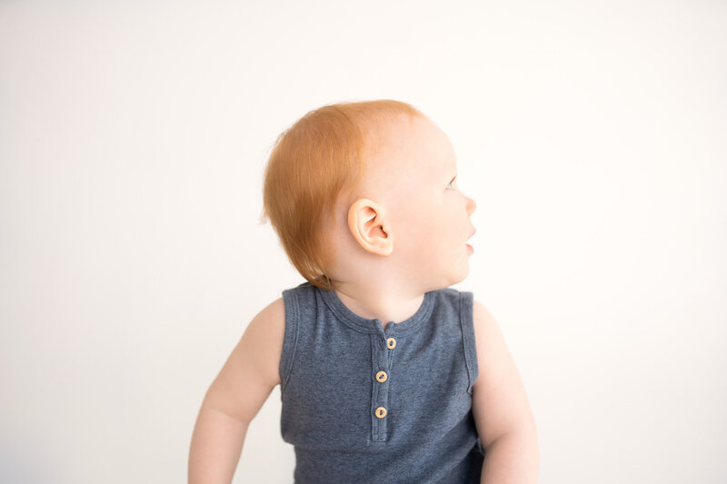 little boy with white backdrop in a teal onsie sitting on a tan blanket and smiling at camera