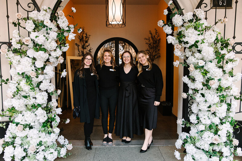 Four SoHo63 team members pose beneath a floral-covered archway at the entrance of SoHo63, a Chandler wedding and event venue. All are dressed in black and smiling, standing in front of the venue’s signature arched doors framed by lush white blooms and greenery.