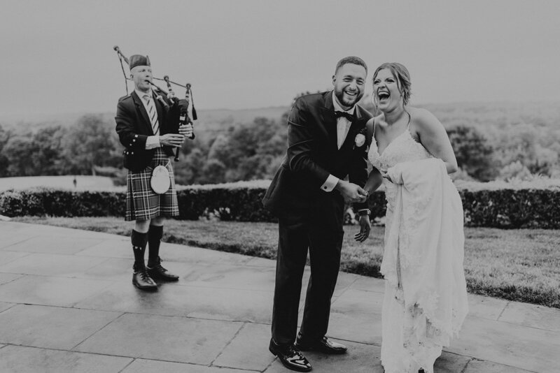 Bride and Groom laughing together, holding hands while dancing in front of irish bagpiper, with rolling hills in the background.