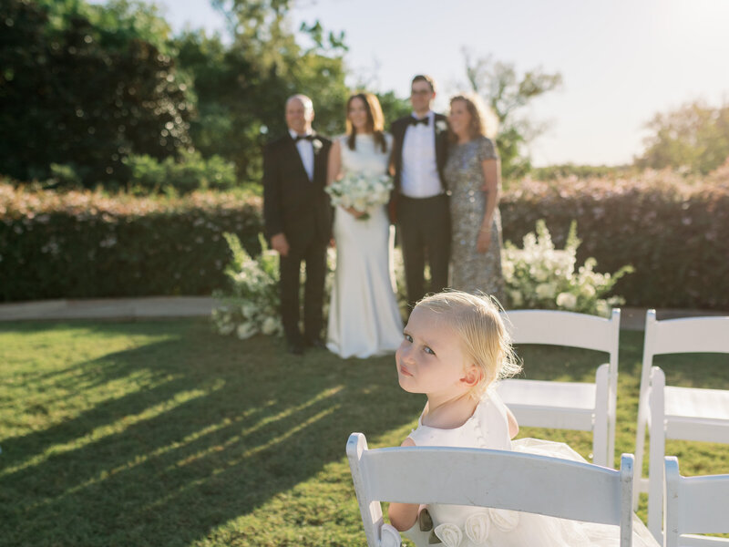 Flower girl sitting in a ceremony chair looking at the camera while bride poses for a family photo in the background