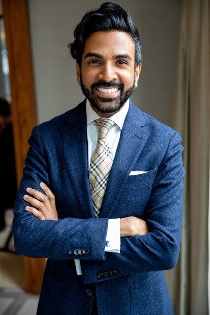 Headshot of Shaun Rajah with navy suit, Burberry classic check tie and very well tailored suite. Beard and suave hair. 