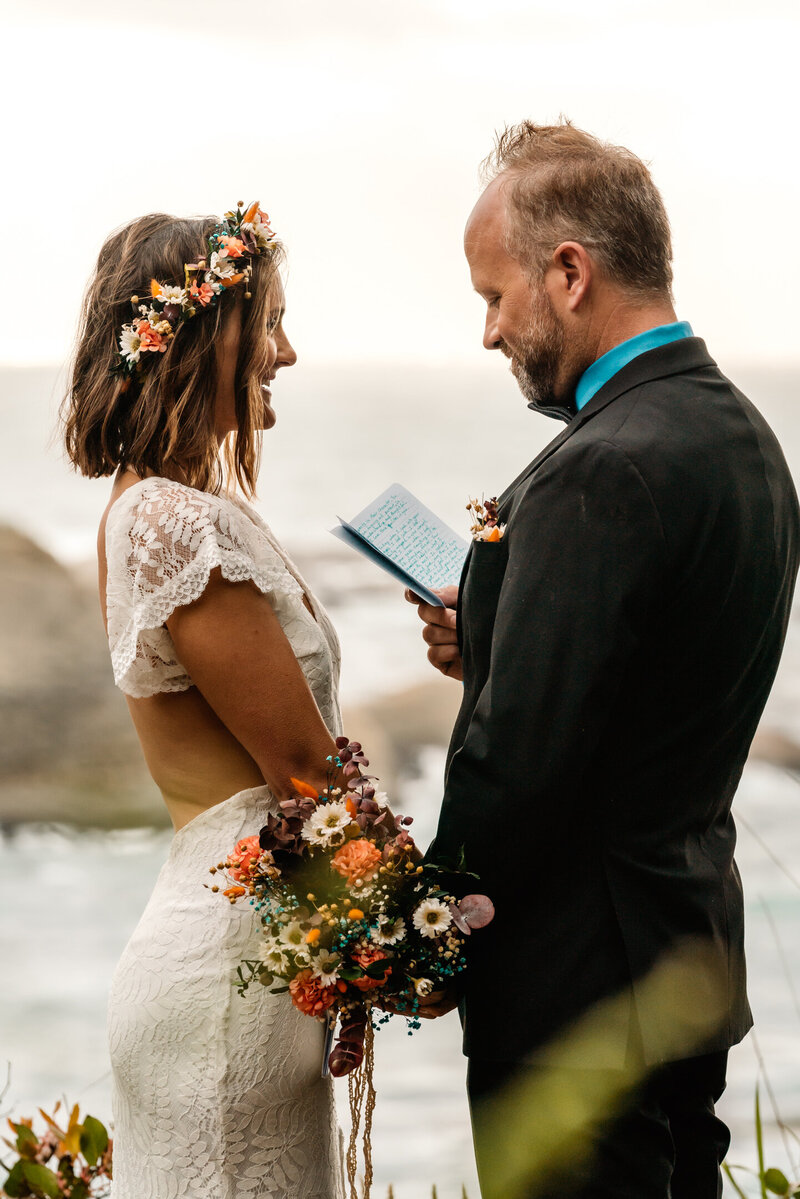 during their coastal elopement, a bride annd groom hold hands and smile widely at each other. They exchange vows standing above the sea