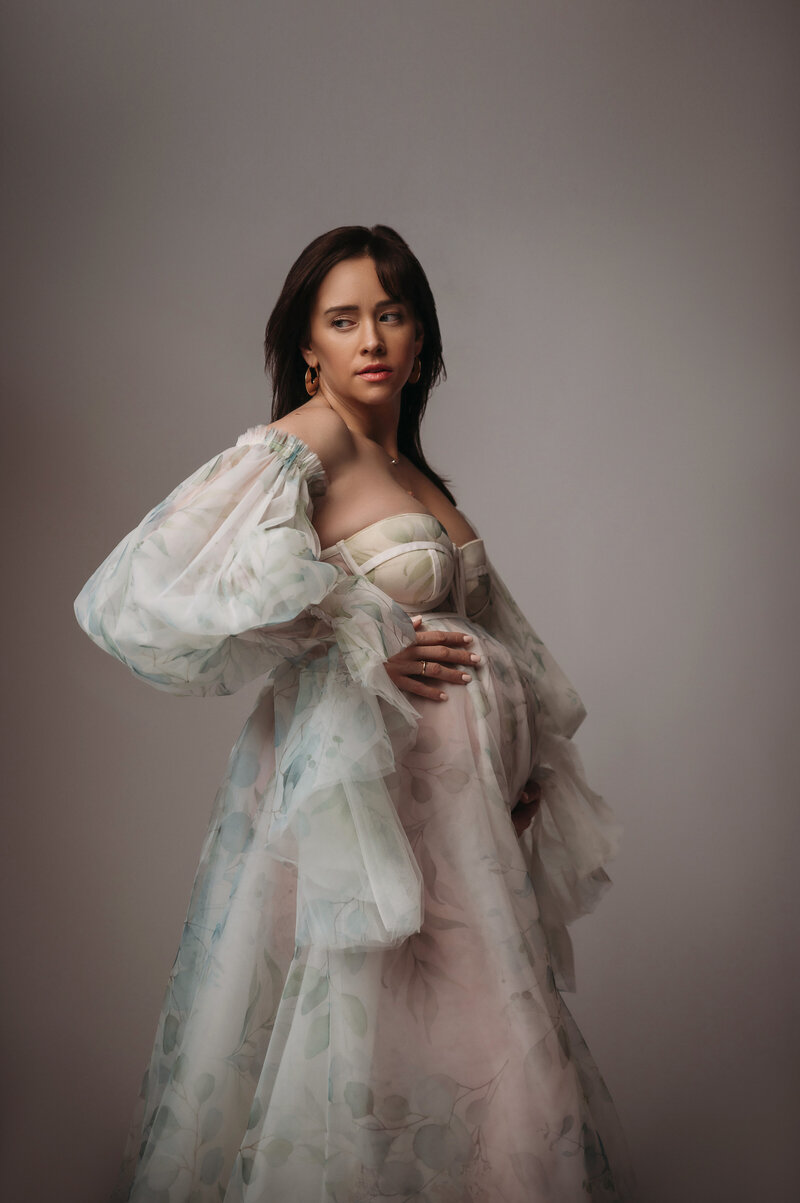 beautiful pregnant woman with brown hair wearing whimsical sage green and white gown while holding her baby bump