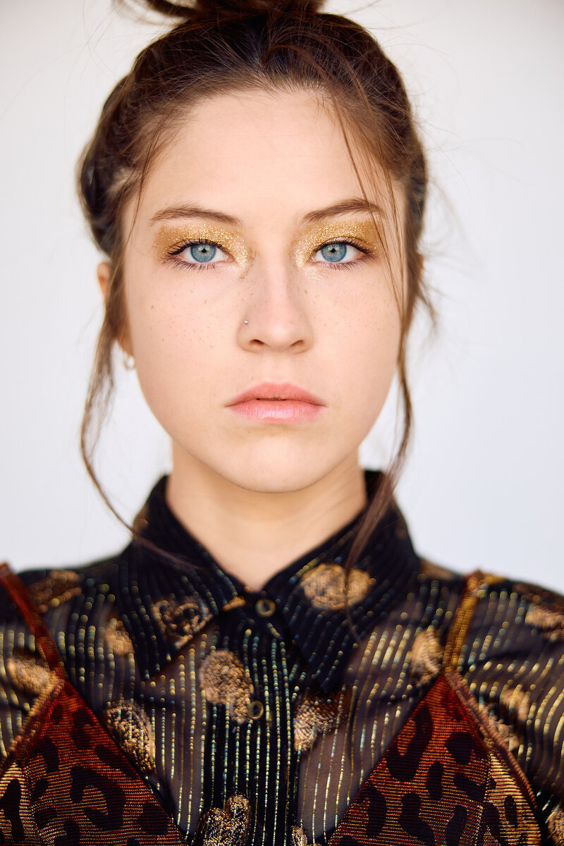 Striking close-up of a young woman with bright blue eyes and gold glitter eyeshadow, wearing a sheer black and gold patterned top. Her hair is pulled back, highlighting her delicate features and piercing gaze.