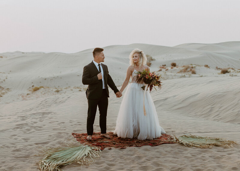 Bride and groom holding hands while standing on a carpet with palm leaves on top of the sand dunes at golden hour in Utah.