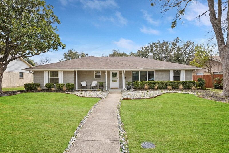 Single-story ranch-style home with a wide front lawn, mature trees, a covered porch, and updated landscaping in a suburban neighborhood.