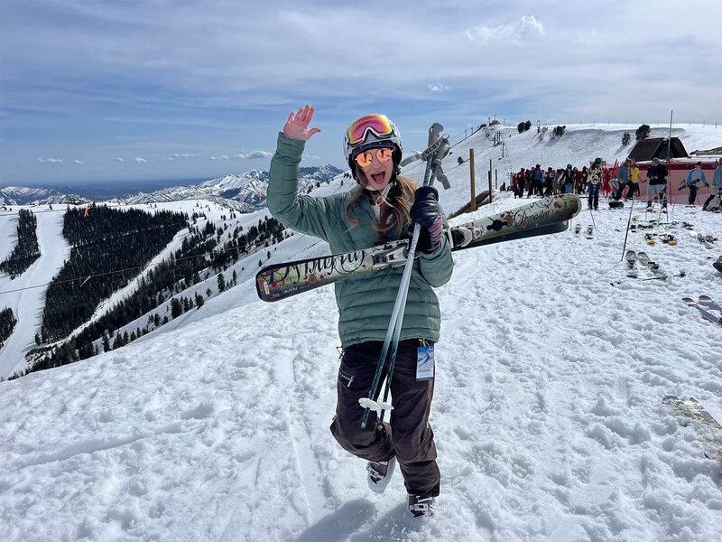 A person wearing ski gear and goggles stands on a snowy slope, holding skis while waving and smiling. There are mountains and other skiers in the background. It's a sunny day with a clear, cloudy sky.