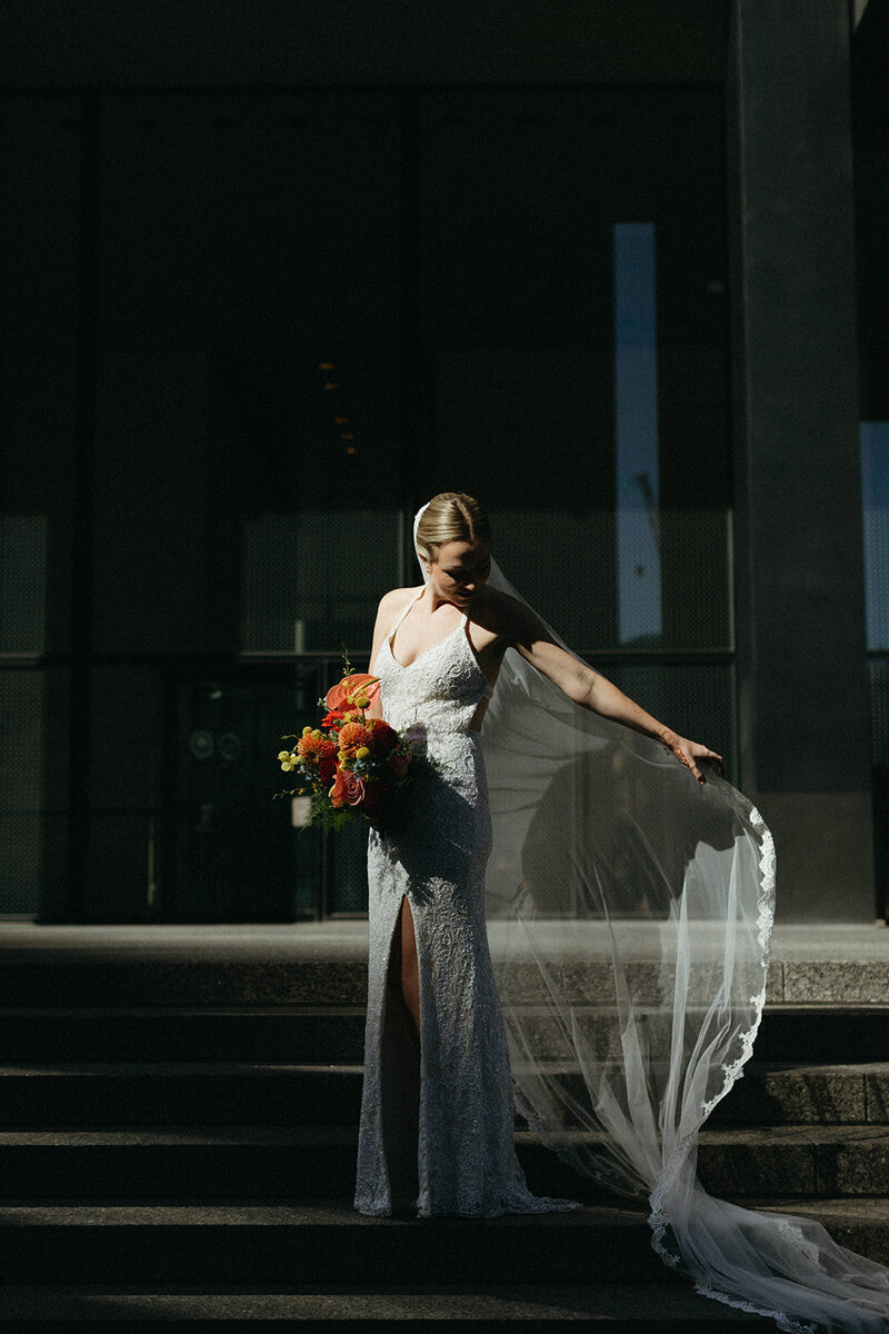 A bride in a lace wedding dress holds a bouquet of vibrant orange flowers, standing gracefully on the steps at Steam Whistle Brewing in Toronto. She gazes down at her veil cascading to the ground, captured beautifully by Toronto wedding photographer, Jennifer van Son..