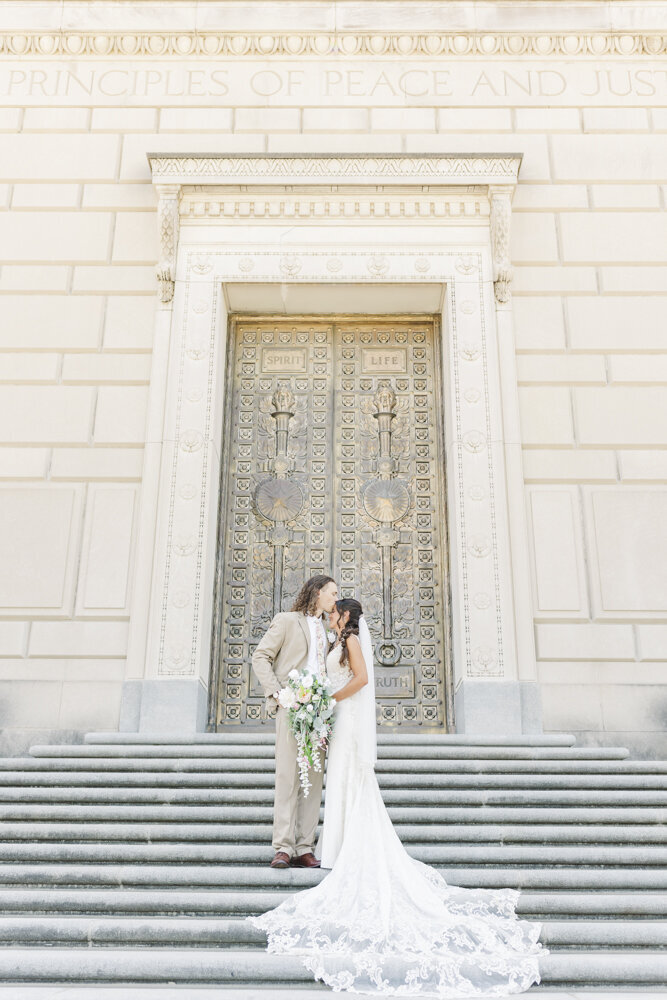 groom kissing bride on the forehead while standing on grand steps in front of a gold door
