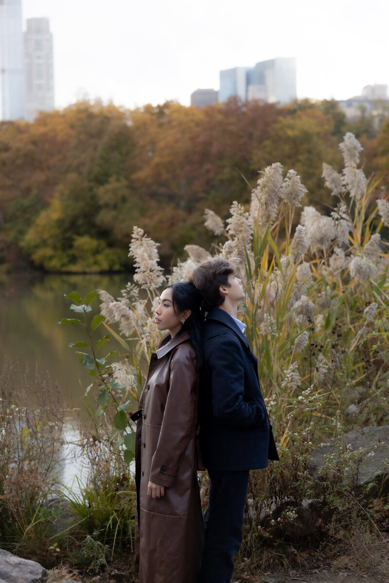 A couple standing back-to-back near Central Park lake in autumn, surrounded by tall grasses and golden foliage, exuding a calm and introspective vibe.