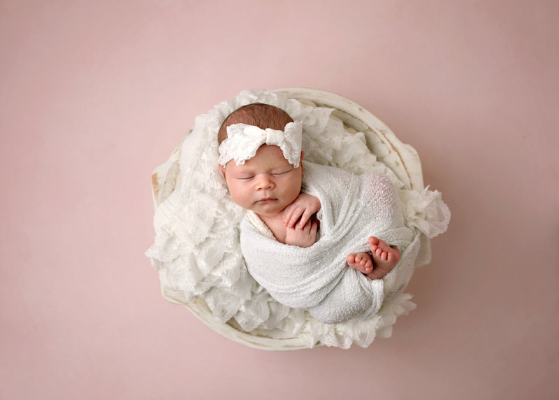 newborn girl with white bow headband wrapped in white cloth  posed in white basket