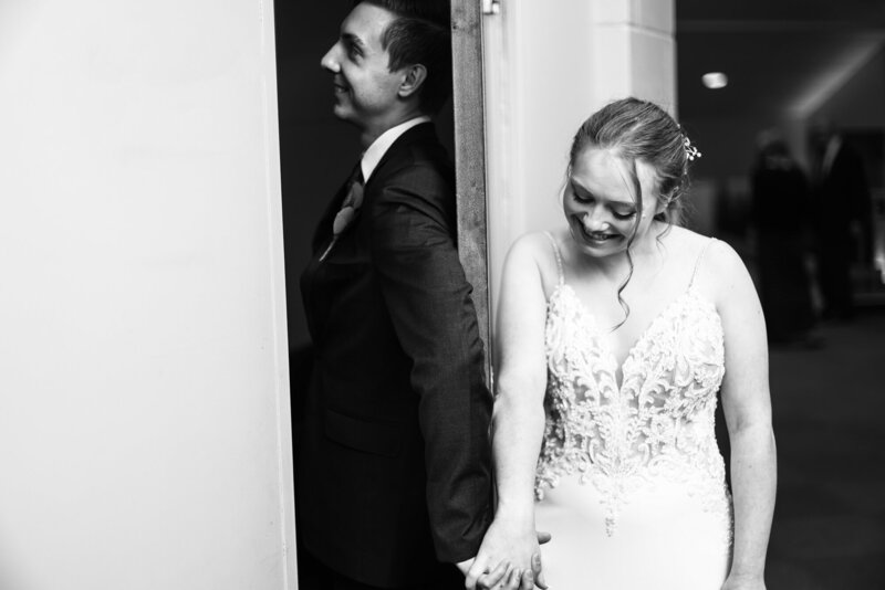 Bride and groom clasp hands on either side of an open door trying not to peek at each other before their wedding ceremony
