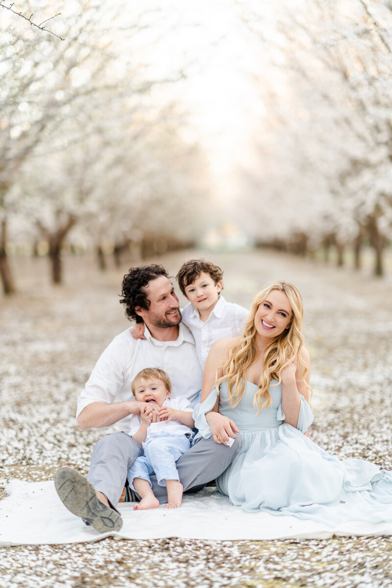 A family of four wearing hues of blues and grays, sit together in a almond blossom field smiling and holding one another, Bay area Family photographer Light Livin Photography.