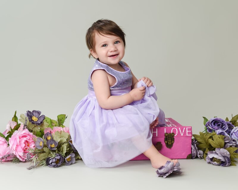 A baby girl sitting on a pink box surrounded by purple and pink flowers, wearing a lavender dress, and smiling sweetly for her first birthday photoshoot.
