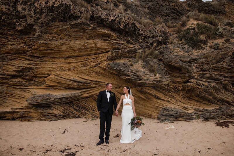 A bride and a groom standing on sand in front of a rocky cliff face