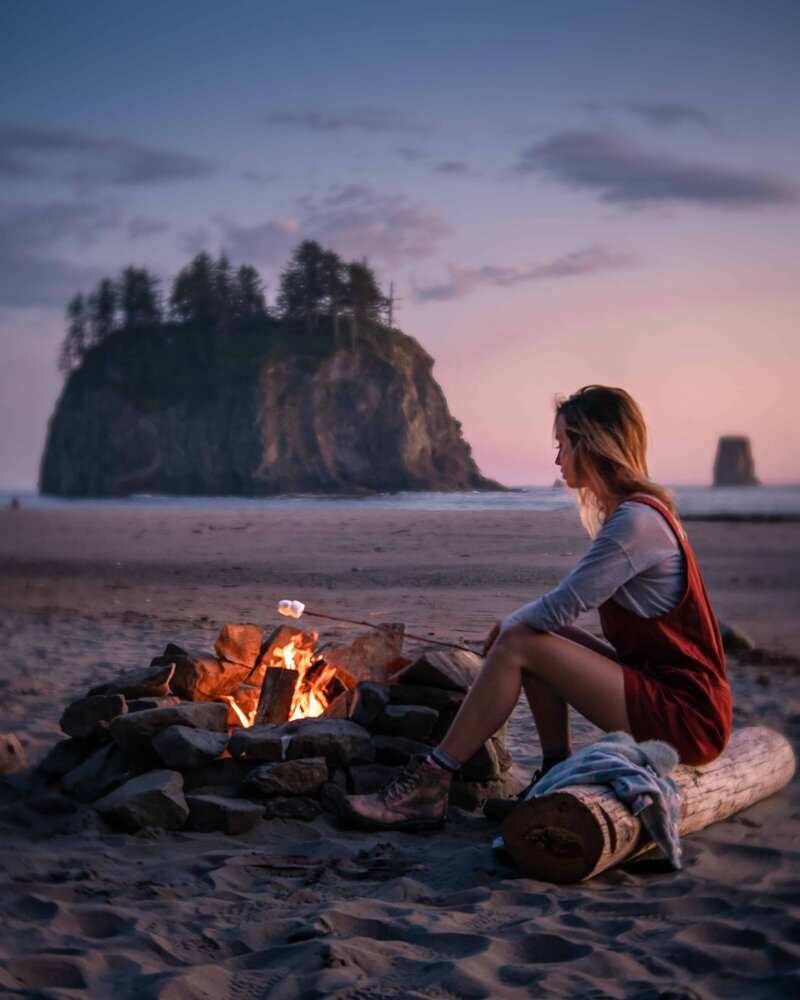 Woman sitting on a log in front of a campfire roasting a marshmallow at sunset with a mountain in the distance
