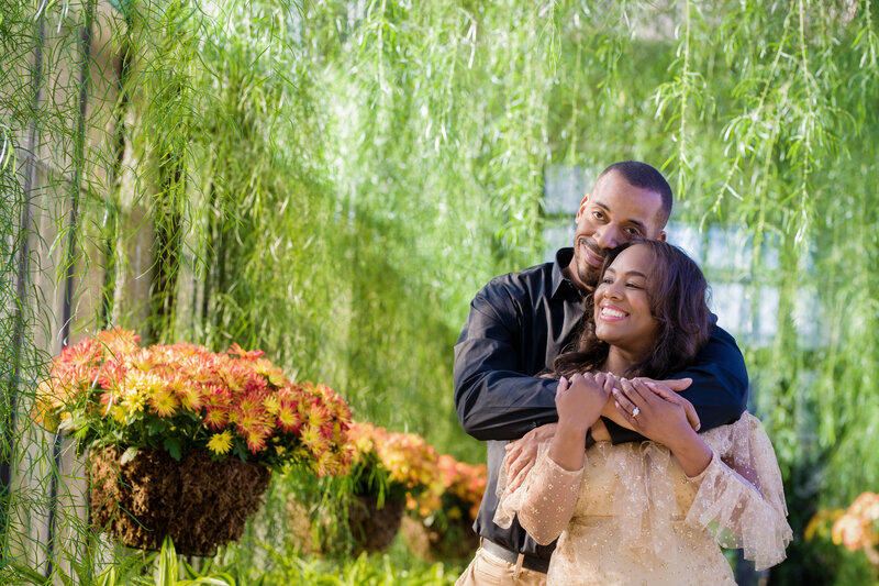 An engaged couple sharing an intimate moment in a lush greenhouse during their engagement session, surrounded by vibrant plants and natural light.