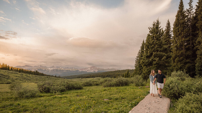 Couple walks on a trail as the sun sets behind the mountains and trees in Vail, Colorado