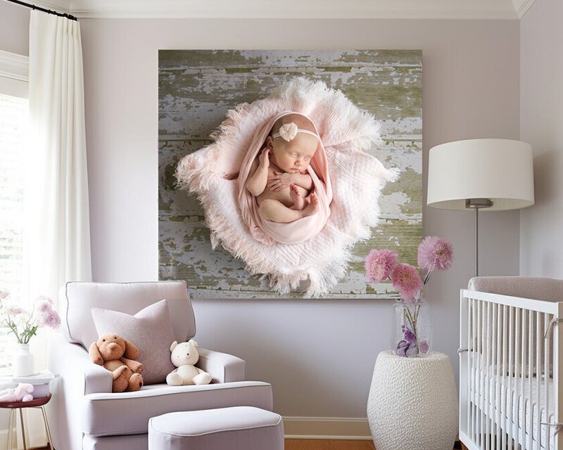 baby girl nursery with lavender arm chair, white crib, lamp and stool, window with light coming in from one side. main focus is the art piece in the center of a beautiful newborn baby girl portrait wrapped in pink