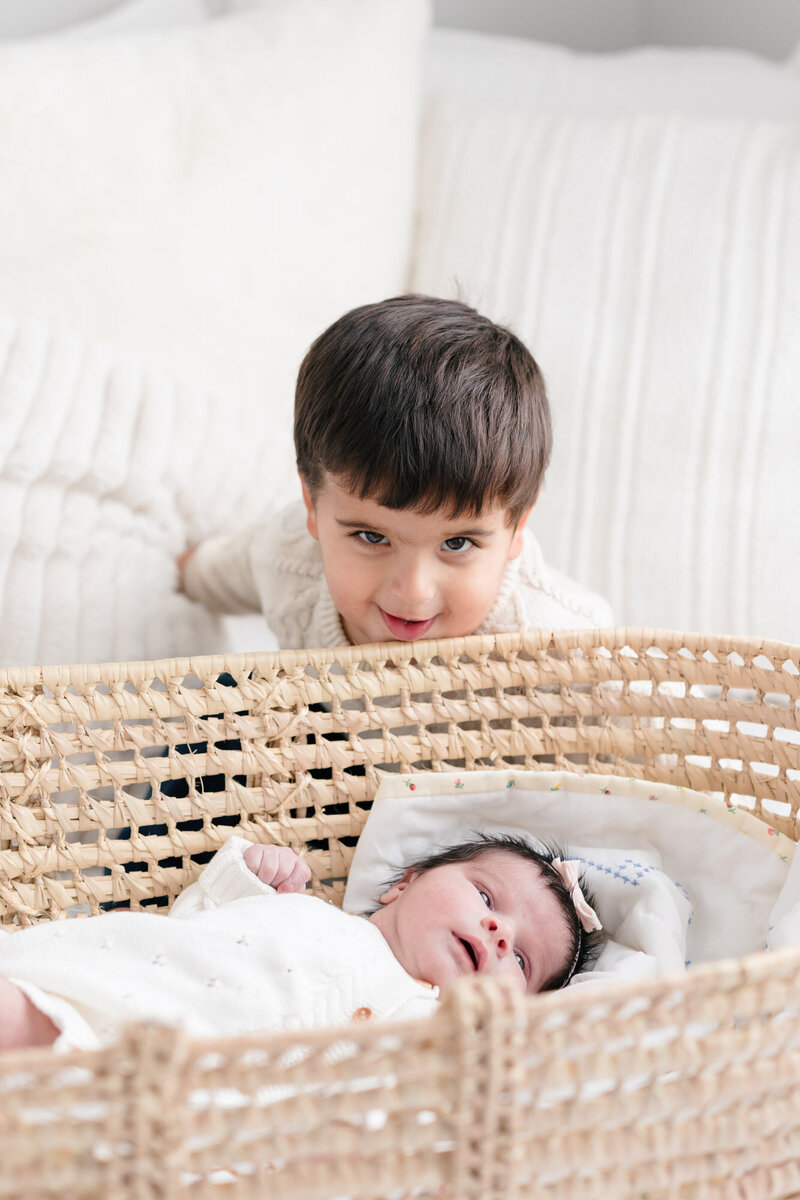 Toddler boy smiling over his baby sister who is laying in a wicker basket