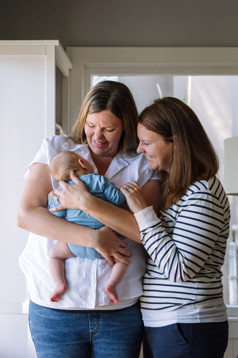 one mom holding newborn as the other mother looks on as they both look at and smile at baby