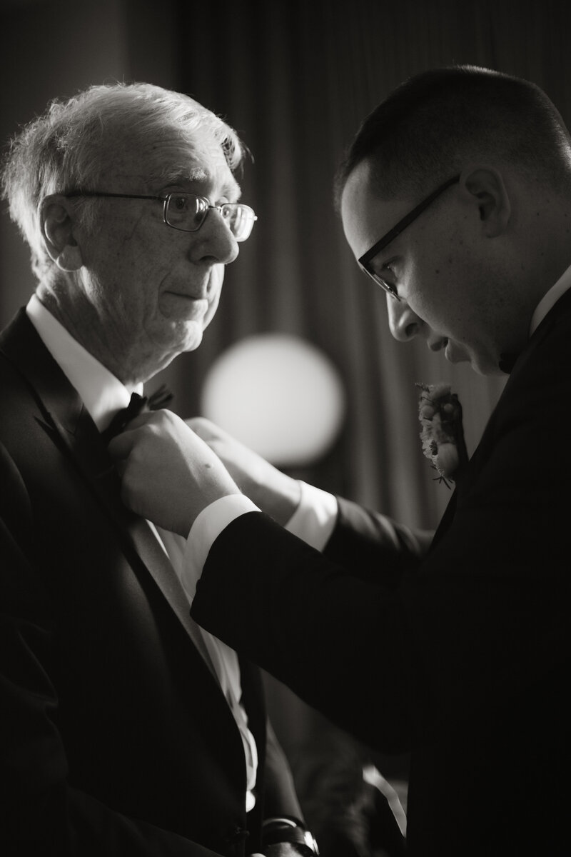 Emotive black and white photo of a groom fixing his father’s bow tie before the ceremony, captured by Daniel from Rainstorm Photo & Video in a candid documentary style.