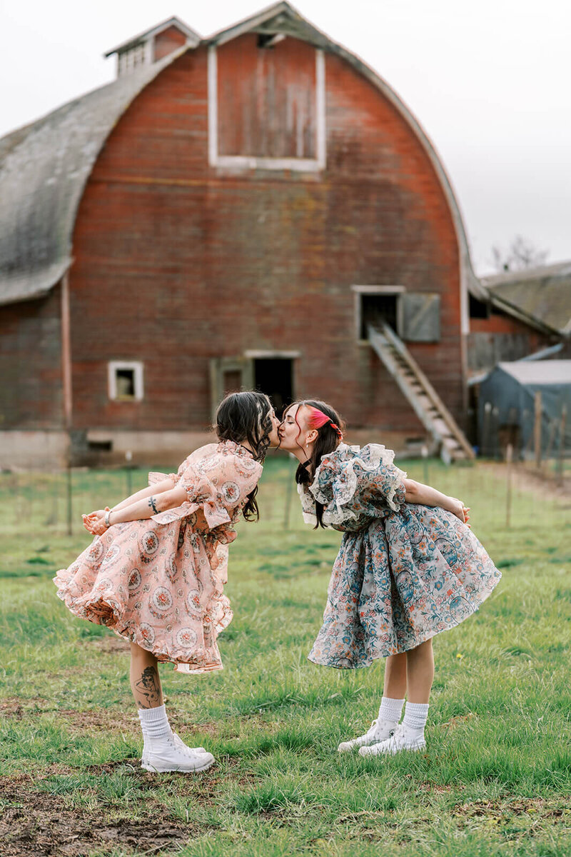 A couple wearing coordinated dresses kiss in front of a barn, representing a styled photoshoot in Oregon. 