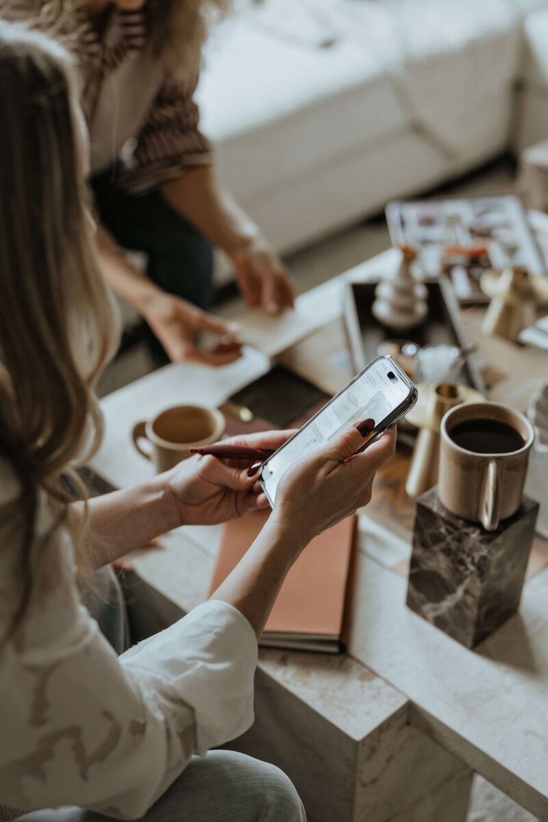 Two interior designers collaborating—one searching for inspiration on her phone as the other reviews fabric samples.