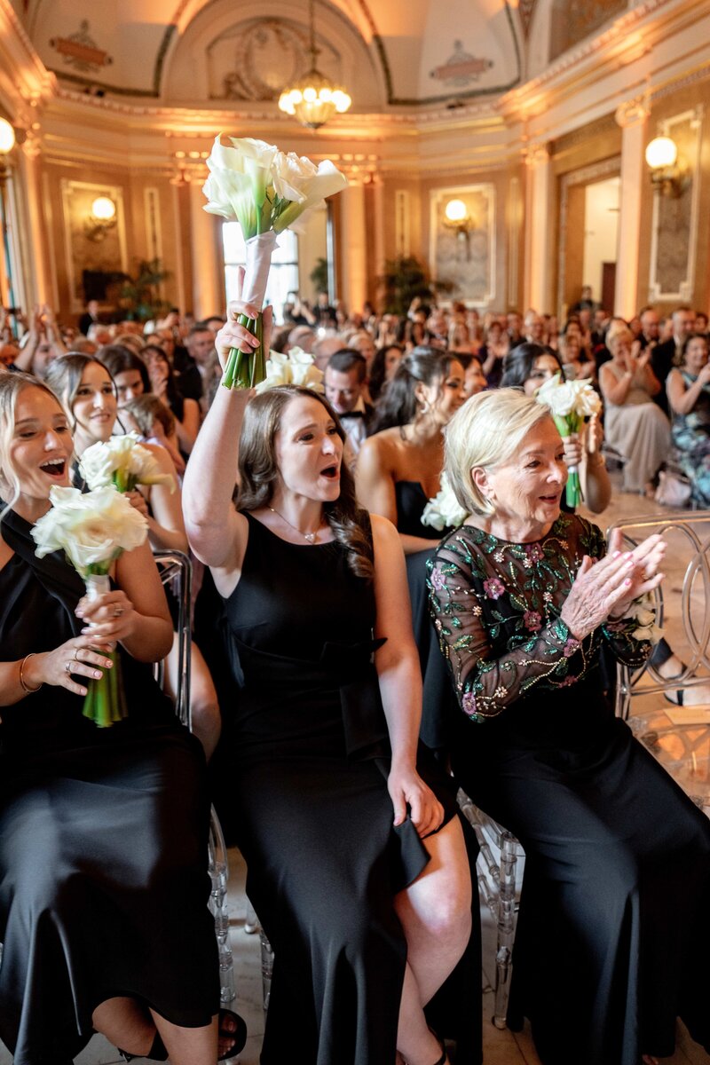 A group of elegantly dressed people at a formal event. A woman in the front row raises a bouquet of flowers excitedly, surrounded by smiling attendees. The room is ornately decorated with warm lighting and intricate architectural details.