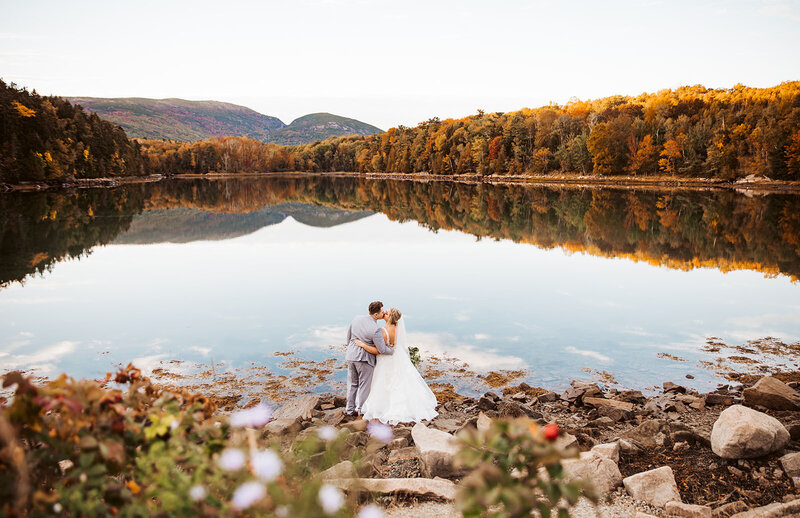 A couple eloping in acadia national park with their backs toward the camera kissing in the fall in front of a reflected lake.