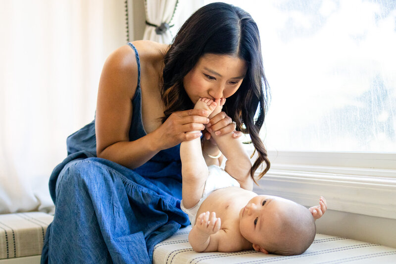 Mother in a blue dress kissing her baby’s feet by a window, tender in-home lifestyle moment captured by Bay Area Photographer Session Info and Pricing - Ellobelle Photography.