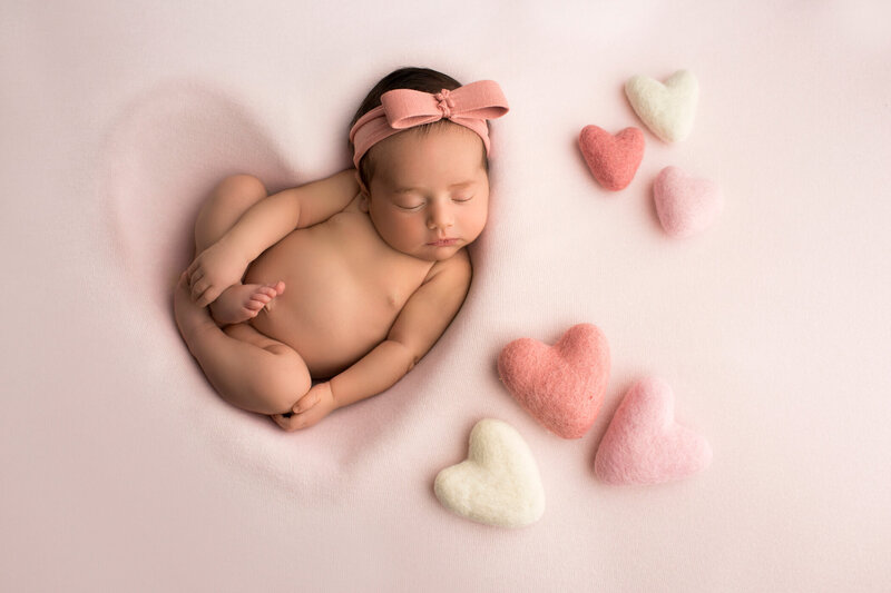 A mother gently cradles her newborn baby against her chest, both looking peaceful and content. The mother has long, straight black hair and is wearing a soft, off-the-shoulder white sweater. The newborn is swaddled in a diaper and has a delicate headband with a floral design. The background is a warm, light brown, adding a cozy and intimate feel to the portrait. The mother's serene expression and the baby's relaxed pose convey a sense of love and tenderness.