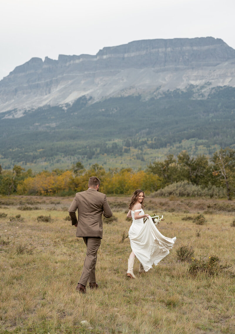 A bride smiles over her shoulder while running through an open Montana field with her groom, surrounded by mountain cliffs and early fall colors, captured by Sydney Breann Photography during their Glacier National Park elopement.