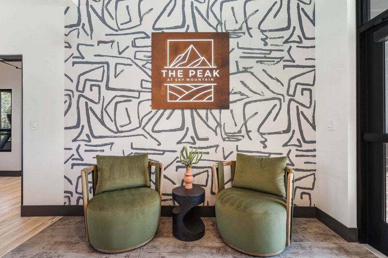 A modern seating area with olive green chairs, a sculptural black table, and a glowing "The Peak at Sky Mountain" sign against bold patterned wallpaper.