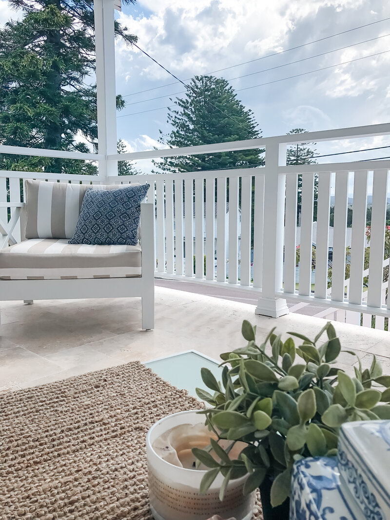 A cozy porch with a white bench, decorative pillows, and a potted plant offers a serene escape. Enjoy the tranquil view of trees under a cloudy sky—courtesy of Newcastle Interior Designer expertise.