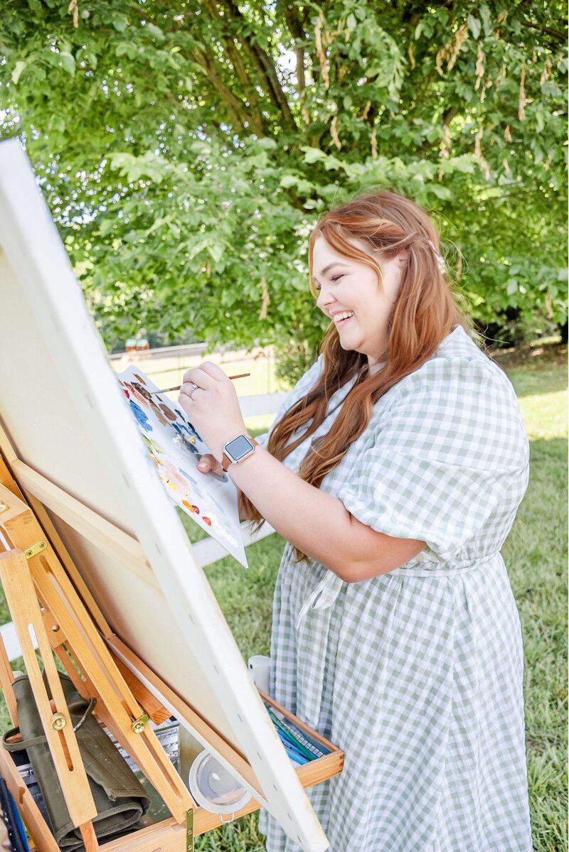 Artist Elyse Stokes creating a live wedding painting, smiling and holding a brush and palette at her easel