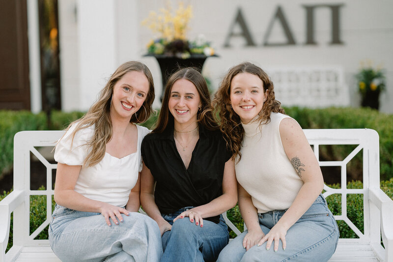 Three young women sitting on a white bench in front of the ADPi house in Lawrence Kansas